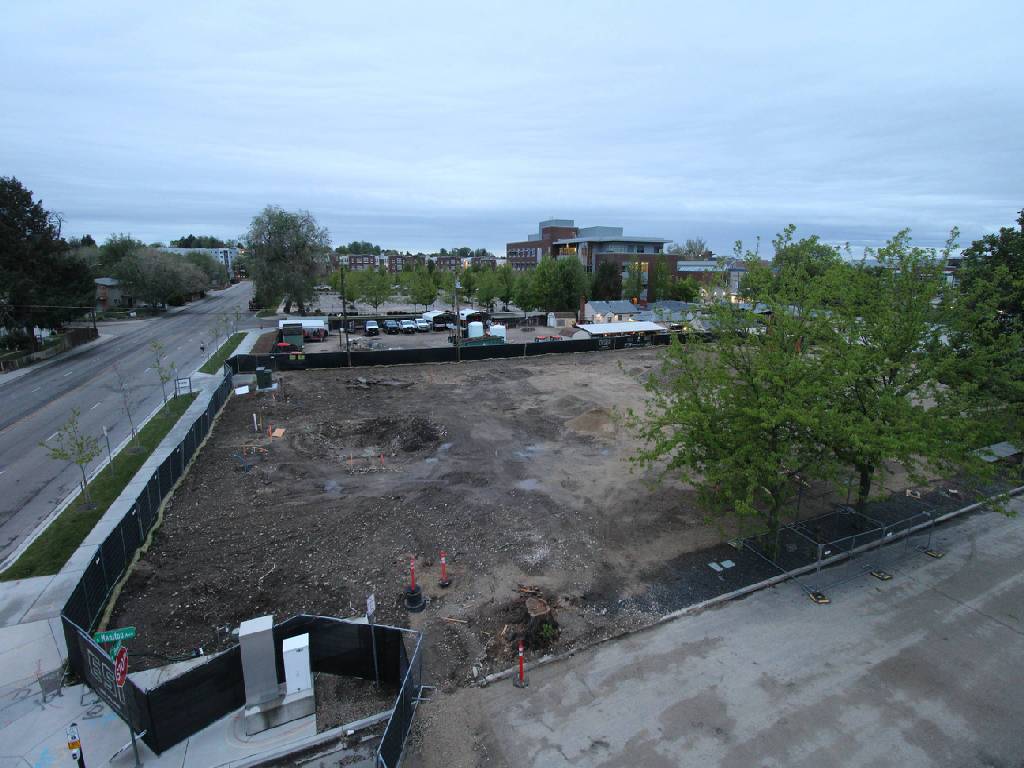 An aerial view shows a vacant, dirt lot surrounded by black fencing in an urban setting. A paved street runs along the left side, with parked cars visible in a lot beyond the vacant area. Buildings and trees are scattered in the background under a clear blue sky. Construction markers are present within the dirt lot.